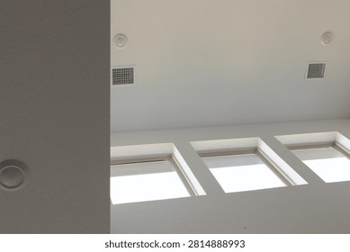 A low-angle view of a modern white ceiling featuring recessed circular lights, square air conditioning vents, and a row of three bright skylights allowing abundant natural sunlight.
