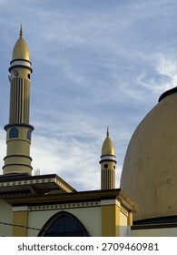 A low-angle view of a mosque featuring tall golden minarets and a large dome against a blue sky with soft clouds. The architectural details combine yellow, gold, and brown tones, showcasing a traditio