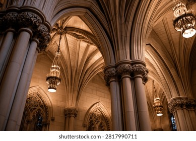 A low-angle view of ornate Gothic architecture, featuring ribbed vaults, carved columns, and hanging chandeliers within a majestic cathedral.