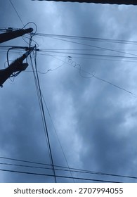 A low-angle view of overhead power lines and utility poles intersecting against a cloudy sky. The image captures the complexity of urban electrical infrastructure with dramatic weather conditions,