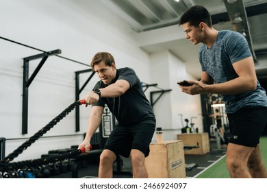 Low-angle view of personal trainer coaching beginner sportsman to perform CrossFit battle ropes exercise in modern fitness gym. Concept of workout training coach business, healthy sports life. - Powered by Shutterstock