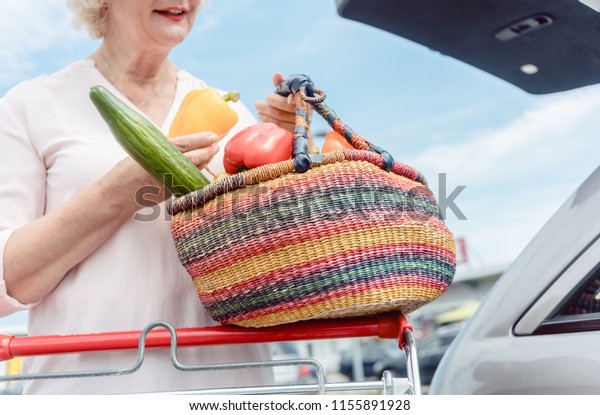 Low-angle view portrait of a cheerful\
senior woman holding a straw basket full of fresh and nutritious\
vegetables after shopping session at the\
hypermarket