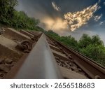 Low-angle view of a railway track stretching into the horizon, surrounded by greenery, with dramatic sunset clouds glowing in the sky, capturing a sense of journey and solitude.