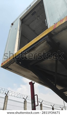 Low-angle view of a raised hydraulic loading dock with an open refrigerated truck trailer at a warehouse logistics facility. Industrial cargo handling equipment used for loading and unloading goods in the transportation and cold chain distribution industry under a clear blue sky.