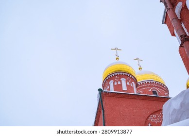 A low-angle view of a red brick Orthodox church with two bright golden onion domes topped with crosses, set against a vast pale blue winter sky