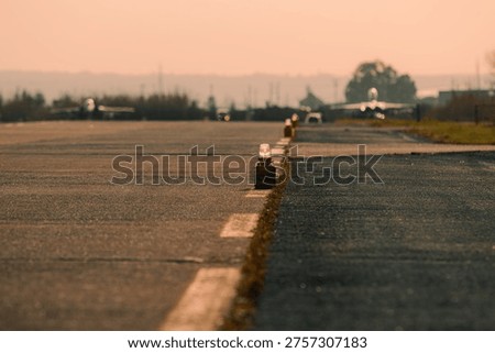 Low-angle view of runway edge lights aligned along an airfield at sunset, with shallow depth of field and warm golden tones. The foreground lighting units are in sharp focus while the background, including aircraft and airport infrastructure, remains softly blurred. Atmospheric heat haze and cinematic lighting create a strong aviation mood.