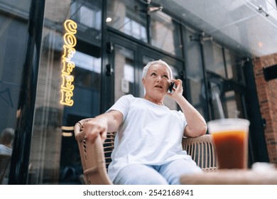 Low-angle view of serious senior woman using smartphone sitting in comfortable chair outside coffee shop, enjoying refreshing iced coffee. Elderly woman relaxing in chair outside cafe. - Powered by Shutterstock