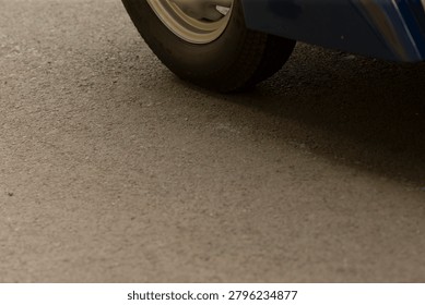 Low-angle view showing the lower edge of a blue vehicle and a single front wheel resting on coarse textured asphalt. Soft natural lighting highlights tire