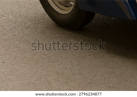 Low-angle view showing the lower edge of a blue vehicle and a single front wheel resting on coarse textured asphalt. Soft natural lighting highlights tire
