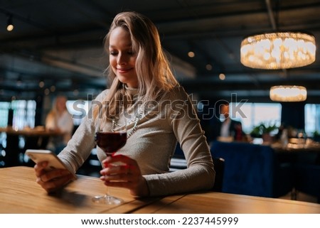 Low-angle view of smiling blonde young woman using smartphone, typing online message sitting at table holding in hand glass of red wine at restaurant. Happy pretty lady chatting with boyfriend.