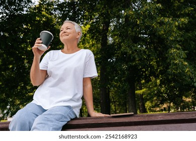 Low-angle view of smiling gray-haired middle-aged woman sipping coffee sitting on bench at city park. Happy serene 60s female resting alone enjoying summertime. Calm pensioner drinking tea outdoors. - Powered by Shutterstock