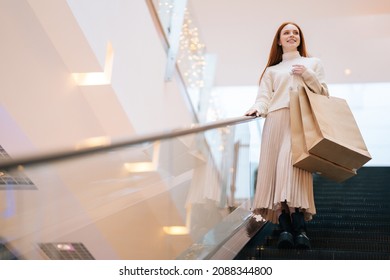 Low-angle view of smiling young woman shopper holding on escalator handrail and riding escalator going down in shopping mall, looking away, paper bags with purchases in hands, blurred background.