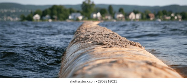 A low-angle view of a stone causeway or path stretching across a body of water, with gentle waves. A small village with houses and trees is visible on the far shore.