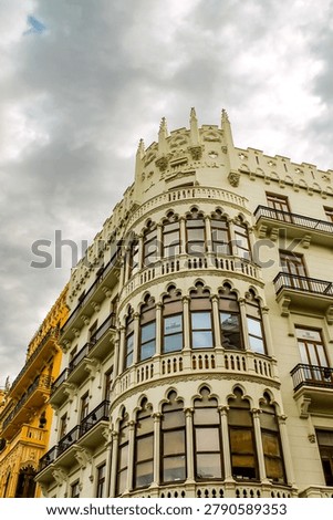 ​A low-angle view of the striking corner facade of the Edificio Equitativa in Valencia, Spain. The image highlights the detailed Gothic revival architecture, featuring ornate arched windows, balconies, and decorative stonework against a dramatic, cloudy sky.