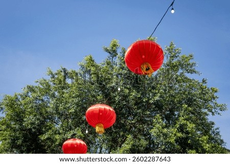 A low-angle view of three bright red lanterns and light bulbs strung between them, set against green trees and a blue sky.
