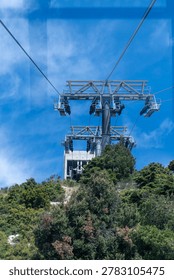 Low-angle view through a window of a cable car approaching heavy steel support towers and upper station on a mountain ridge.
A professional vertical landscape photograph capturing the mechanical engineering of a cable car system. Shot from inside a moving gondola, the image showcases massive dark metal towers holding heavy cables, leading towards a concrete station on a green forested hill under a vivid blue sky. Subtle reflections on the glass add a realistic sense of perspective.