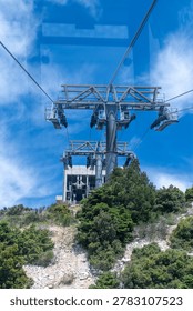 Low-angle view through a window of a cable car approaching heavy steel support towers and upper station on a mountain ridge.
A professional vertical landscape photograph capturing the mechanical engineering of a cable car system. Shot from inside a moving gondola, the image showcases massive dark metal towers holding heavy cables, leading towards a concrete station on a green forested hill under a vivid blue sky. Subtle reflections on the glass add a realistic sense of perspective.