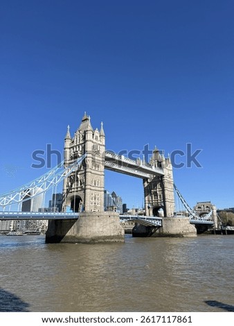 Low-angle view of London’s Tower Bridge on a sunny day, showing Gothic details, blue suspension, River Thames below, and modern skyline in the back—classic meets contemporary