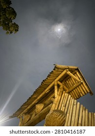 Low-angle view of a traditional bamboo structure under a cloudy night sky with the glowing moon partially hidden behind clouds. Natural bamboo architecture illuminated by warm light creates a peaceful rural atmosphere, blending craftsmanship, sustainability, and nature in an outdoor setting.