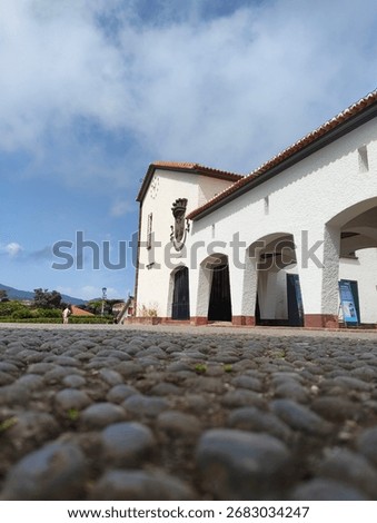Low-angle view of a traditional whitewashed building with cobblestone foreground in Madeira, Portugal, under a blue sky with scattered clouds.
