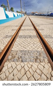 Low-angle view of tram tracks embedded in a cobblestone street, leading towards a distant horizon with blue sky and clouds. The scene is urban with buildings and parked cars visible.