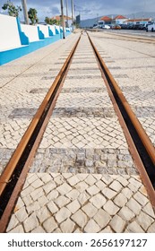 Low-angle view of tram tracks embedded in a cobblestone street, leading towards a distant horizon with blue sky and clouds. The scene is urban with buildings and parked cars visible.