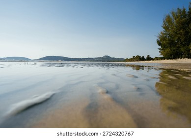 Low-angle view of a tranquil beach with shallow seawater reflecting the blue sky, gentle ripples on wet sand, distant hills on the horizon, and coastal trees creating a peaceful seaside atmosphere.
