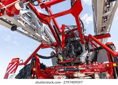 Low-angle view under a large red agricultural sprayer showing the metal chassis, hydraulic components, folding booms, wheels and nozzle assemblies against a