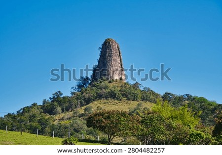 Low-angle view of a unique monumental volcanic rock formation rising from a green forested hill under a clear blue sky in Brazil.