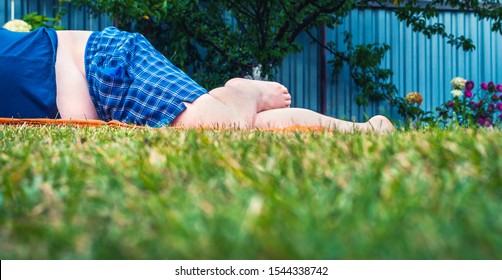 Low-Angle View Of Unrecognizable Barefoot Big Fat Man In Blue Shirt And Plaid Shorts Lying On Mowed Green Grass Lawn With Blurred Garden, Fence Background. Copy Space.