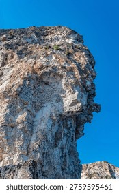 Low-angle view of a weathered limestone cliff with textured rock and clear blue sky, featuring a natural formation that resembles a human side profile.