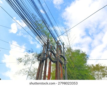 Low-angle view of weathered, rusty utility poles supporting a dense network of black overhead power lines and fiber cables against a vibrant blue sky. Green bamboo foliage provides a natural contrast to the industrial urban clutter.