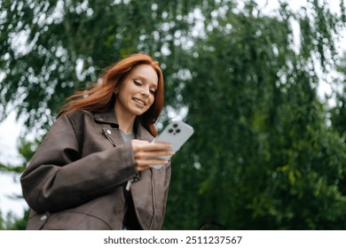 Low-angle view of young woman with long red hair joyfully texting on mobile phone while strolling through city street on summer day. Cheerful redhead female happily messaging on smartphone outdoors. - Powered by Shutterstock