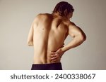 Lower back pains. Rear view of shirtless young man suffering from back pains against grey studio background. Concept of men