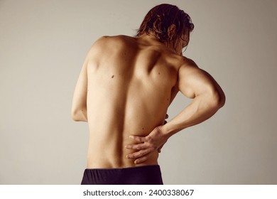 Lower back pains. Rear view of shirtless young man suffering from back pains against grey studio background. Concept of men's beauty, health, body care, sportive lifestyle, medicine