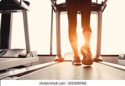 Lower Body At Legs Part Of Fitness Girl Running On Running Machine Or Treadmill In Fitness Gym With Sun Ray. Warm Tone. Healthy And Exercise Activity Concept. Workout And  Strength Training Theme.