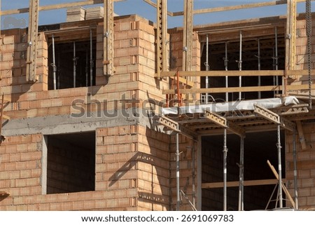 lower facade showing scaffold platforms and open bays, stacked blockwork, metal props and safety railings, gritty textures, closeup perspective suggesting trade work and material staging at ground