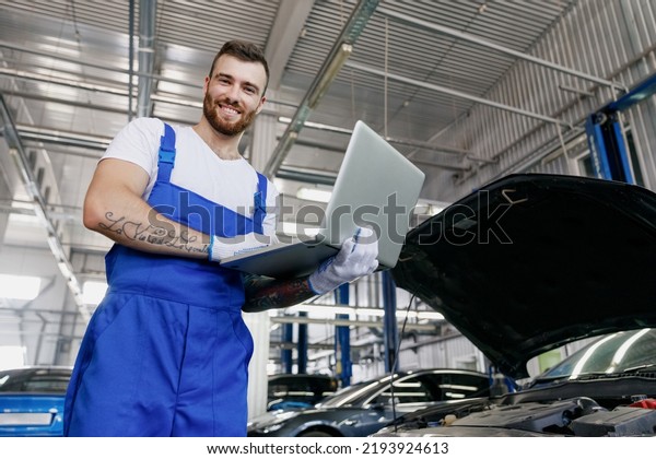 Lower fun young professional technician car\
mechanic man wearing overalls t-shirt use hold laptop pc computer\
make diagnostics fix problem with raised hood work in vehicle\
repair shop workshop\
indoors