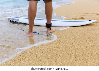 Lower half of surfer on sand, standing on the beach with an attached surfboard near the ocean - Powered by Shutterstock