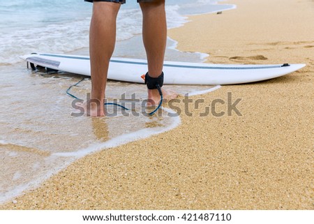 Lower half of surfer on sand, standing on the beach with an attached surfboard near the ocean