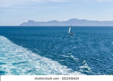 Lower Left Corner Cut By The Wake Of A Large Ship. On The Right A Small Sailboat Approaches The Wake And In The Background Some Bluish Mountains On The Horizon