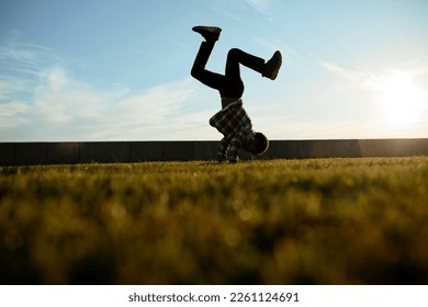 Lower outdoor view of african american teenager training break dance move, balancing on hands with raised legs up, doing cartwheel on green lawn of public park in evening. Urban culture and activity - Powered by Shutterstock