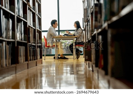 Lower shot through space between bookshelves show Hispanic man sit and discuss with Asian woman at table in public library in front of glass window with day light.