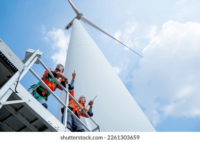 Lower view of two engineers or technician workers stand on base of big windmill or wind turbine and point or look to left side with blue sky.