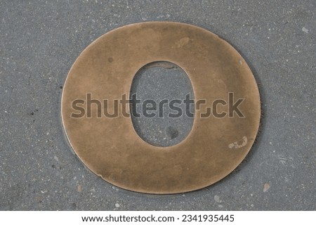 Lowercase letter O in bronze on the asphalt of a street