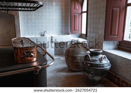 Low-key image of a rustic historical kitchen with a vintage copper pot on an old stove, large antique ceramic jars, white tiled walls, and red wooden windows softly lit by natural light