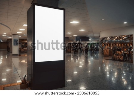 A low-key shot of a vertical blank billboard mock-up stands prominently in the middle of a bustling airport terminal or a shopping mall, with shops in the background, ready for custom advertisements