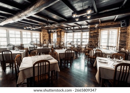 Low-key wide-angle shot of a cozy rustic restaurant interior with wooden beams, stone walls, neatly arranged tables covered in white linens; large bright windows creating an inviting dining atmosphere