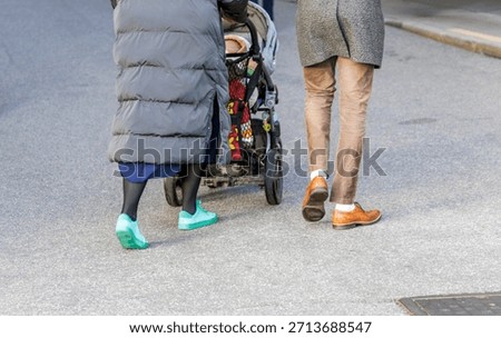 Low-section view of a couple walking on an asphalt street, pushing a baby stroller. Focus on the contrasting footwear.