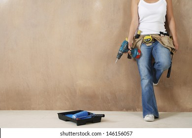 Lowsection of a young woman with toolbelt and drill leaning against wall